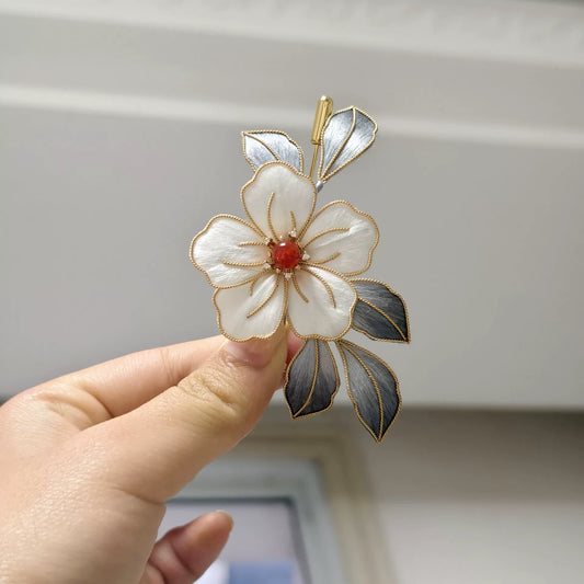 Handheld view of a handmade Ronghua brooch featuring a large white silk velvet flower with gold-toned edges, dark silver-grey leaves, and a red center accent.