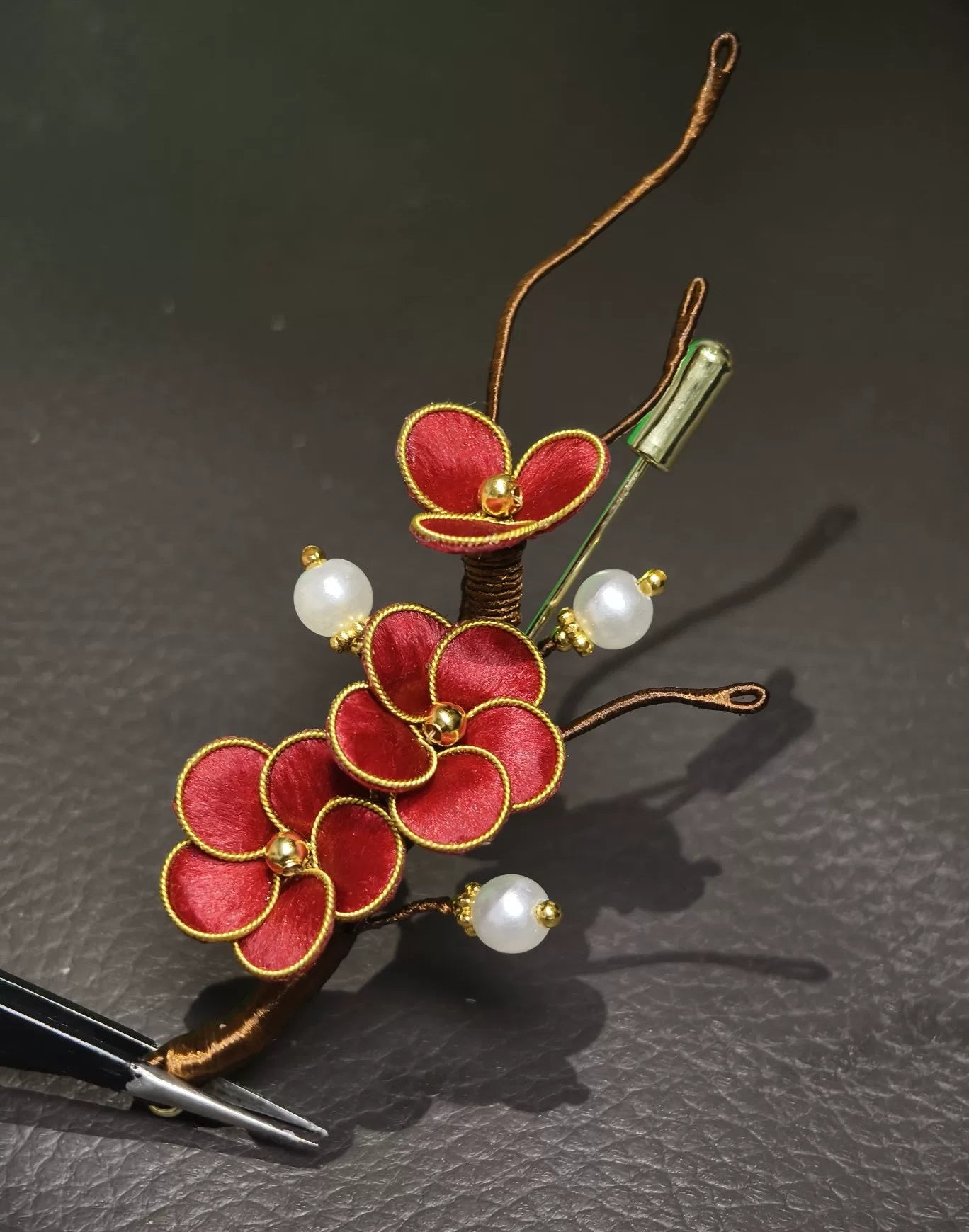 Handmade Ronghua brooch featuring three red silk velvet plum blossoms with gold-toned edges, branching brown stems, and three white pearl accents on a dark surface.