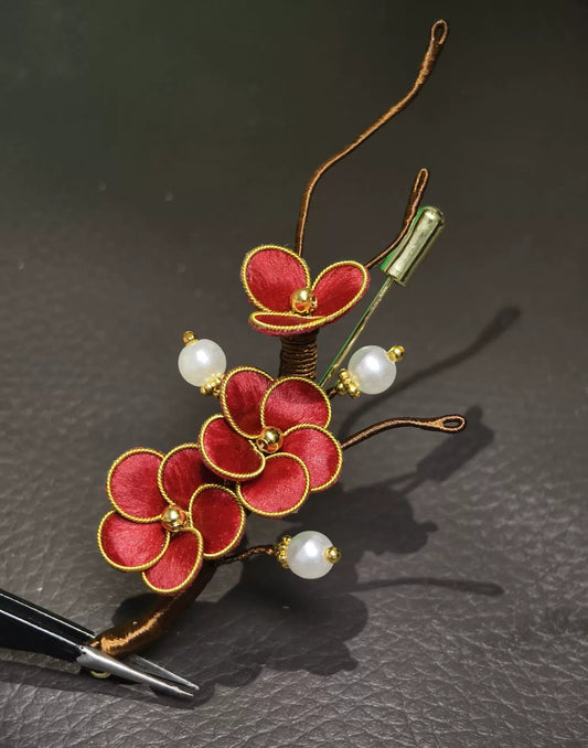 Handmade Ronghua brooch featuring three red silk velvet plum blossoms with gold-toned edges, branching brown stems, and three white pearl accents on a dark surface.