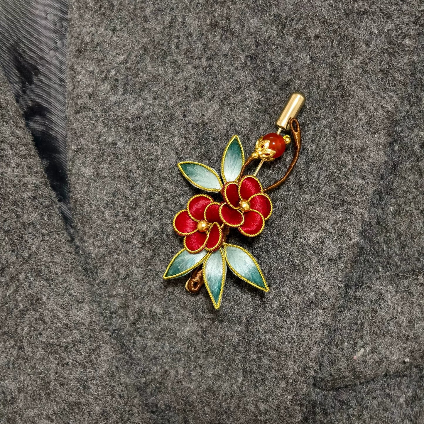 Handmade Ronghua brooch featuring two red silk velvet flowers with gold-toned edges, gradient green leaves, and a red bead accent pinned on grey fabric.