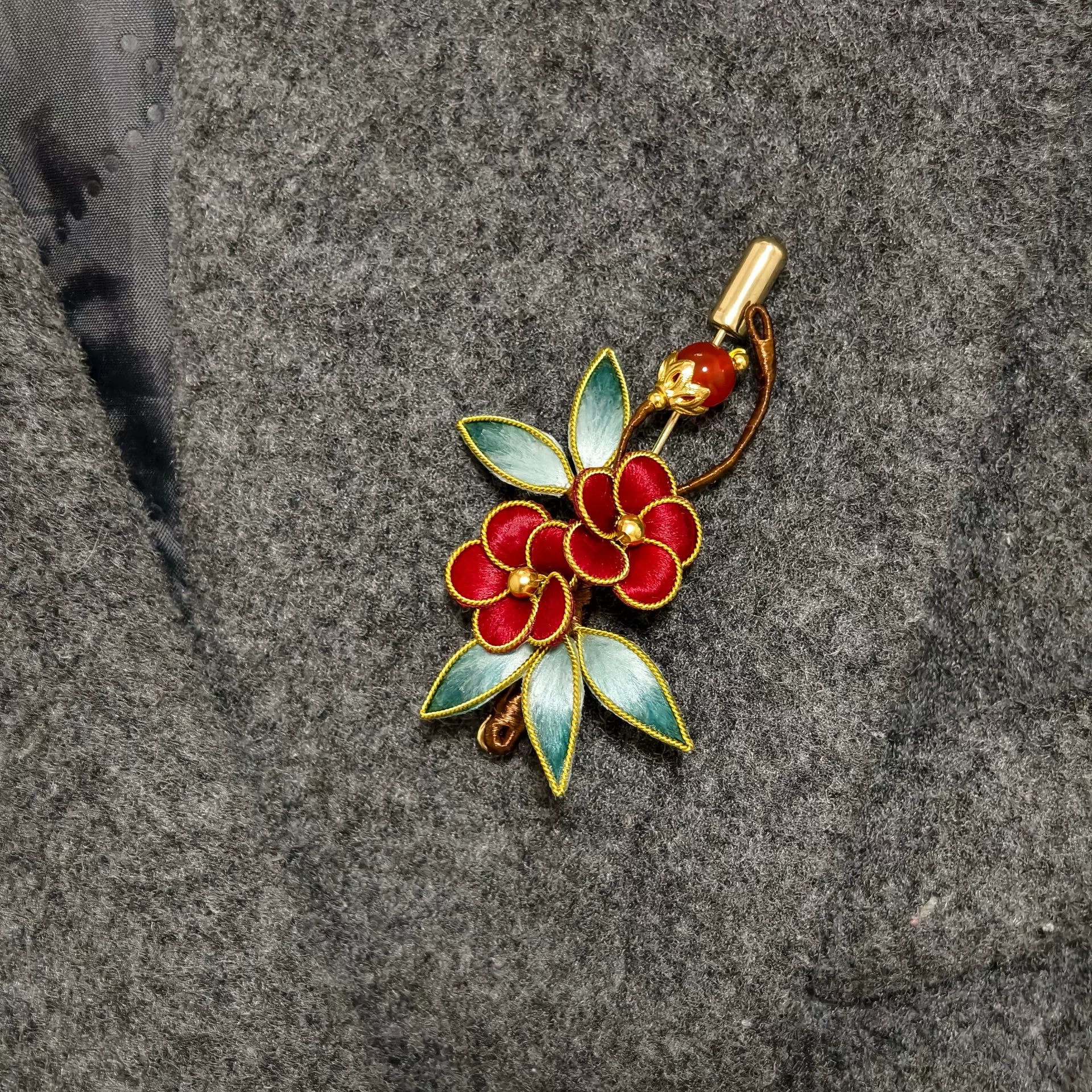 Handmade Ronghua brooch featuring two red silk velvet flowers with gold-toned edges, gradient green leaves, and a red bead accent pinned on grey fabric.