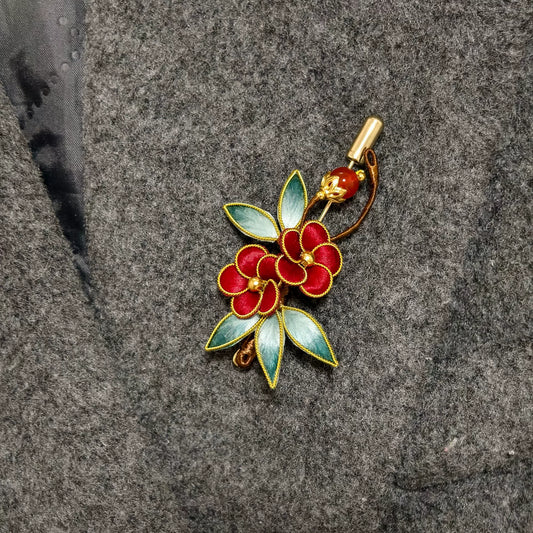 Handmade Ronghua brooch featuring two red silk velvet flowers with gold-toned edges, gradient green leaves, and a red bead accent pinned on grey fabric.