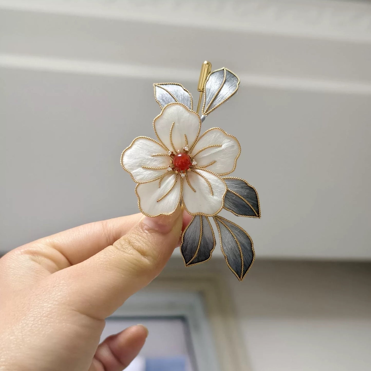 Handheld view of a handmade Ronghua brooch featuring a large white silk velvet flower with gold-toned edges, dark silver-grey leaves, and a red center accent.
