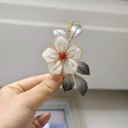 Handheld view of a handmade Ronghua brooch featuring a large white silk velvet flower with gold-toned edges, dark silver-grey leaves, and a red center accent.
