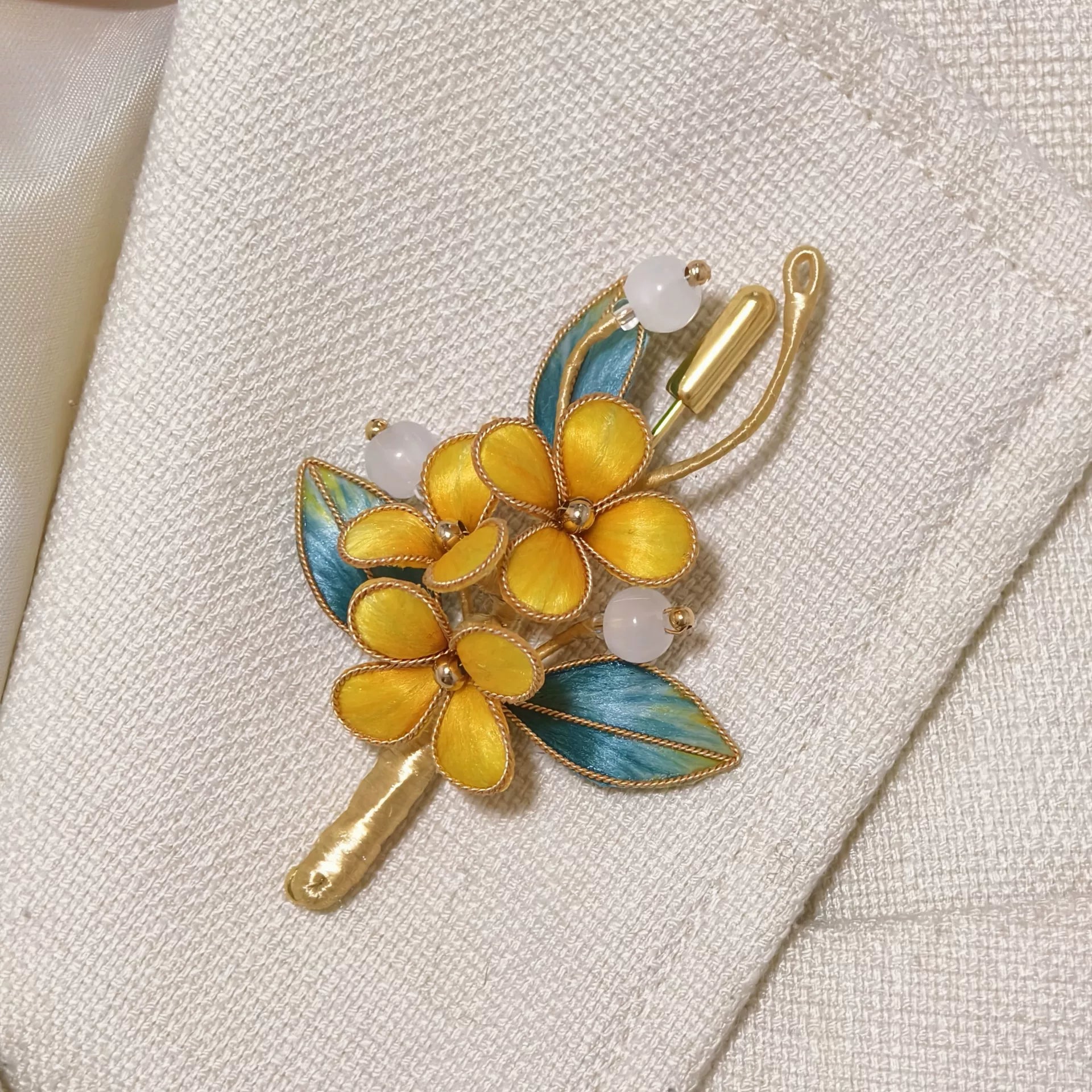 Handmade Ronghua brooch featuring three vibrant yellow silk velvet flowers with gold-toned edges, teal-green leaves, and white beads pinned on textured off-white fabric.