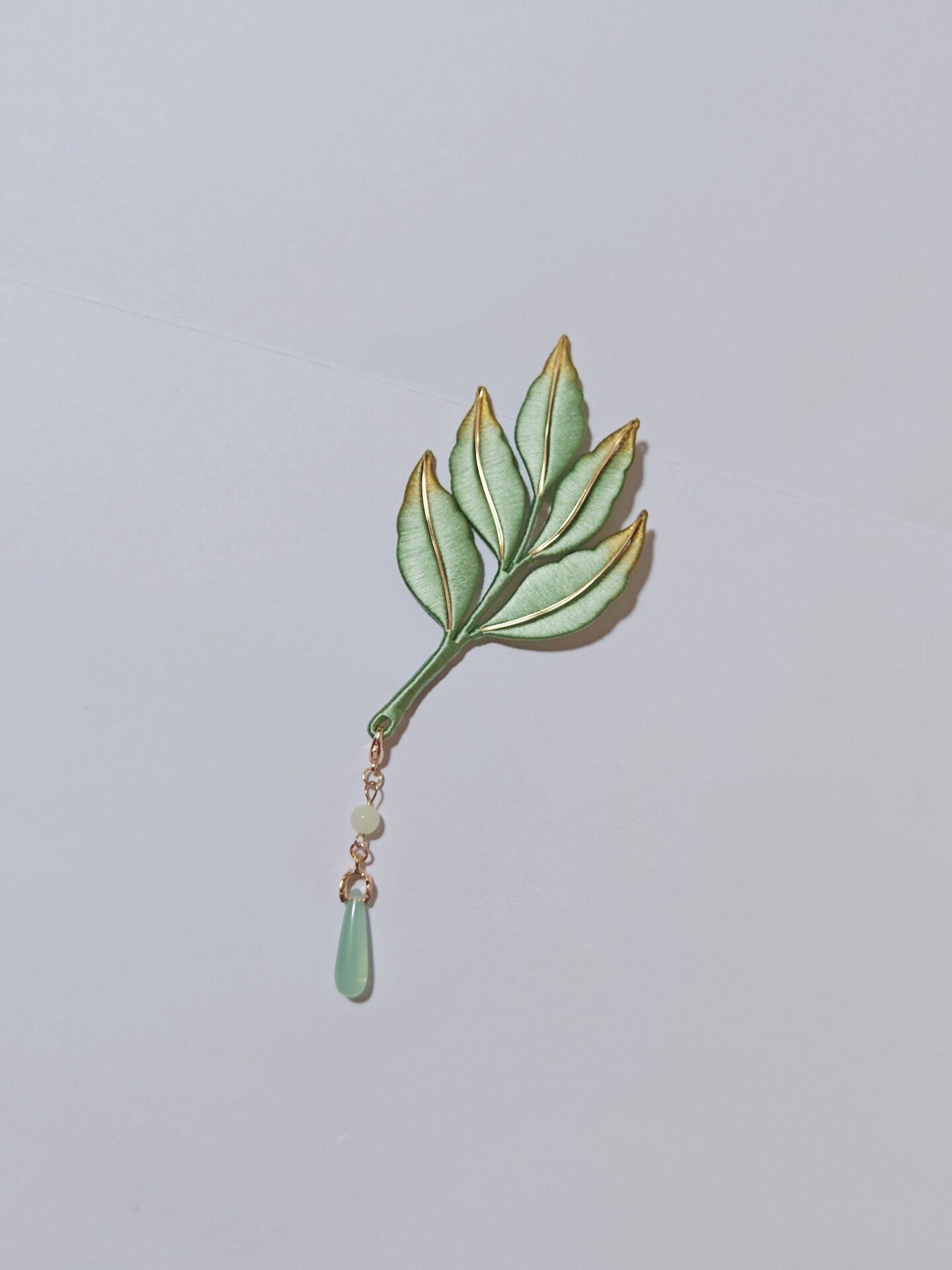 Chanhua handmade green leaf brooch with gold-trimmed edges and jade-green drop pendant on a gold chain, displayed on a light gray background