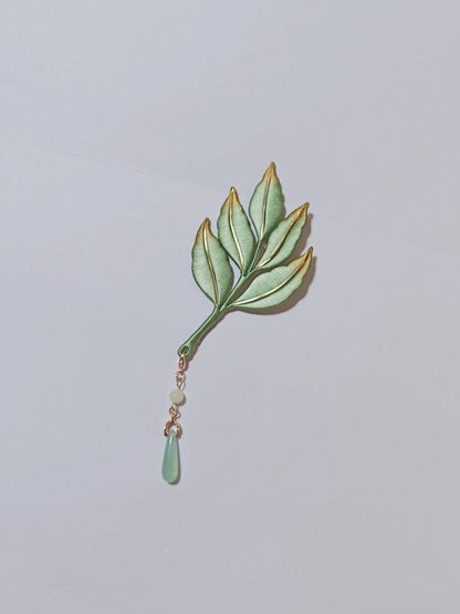 Chanhua handmade green leaf brooch with gold-trimmed edges and jade-green drop pendant on a gold chain, displayed on a light gray background
