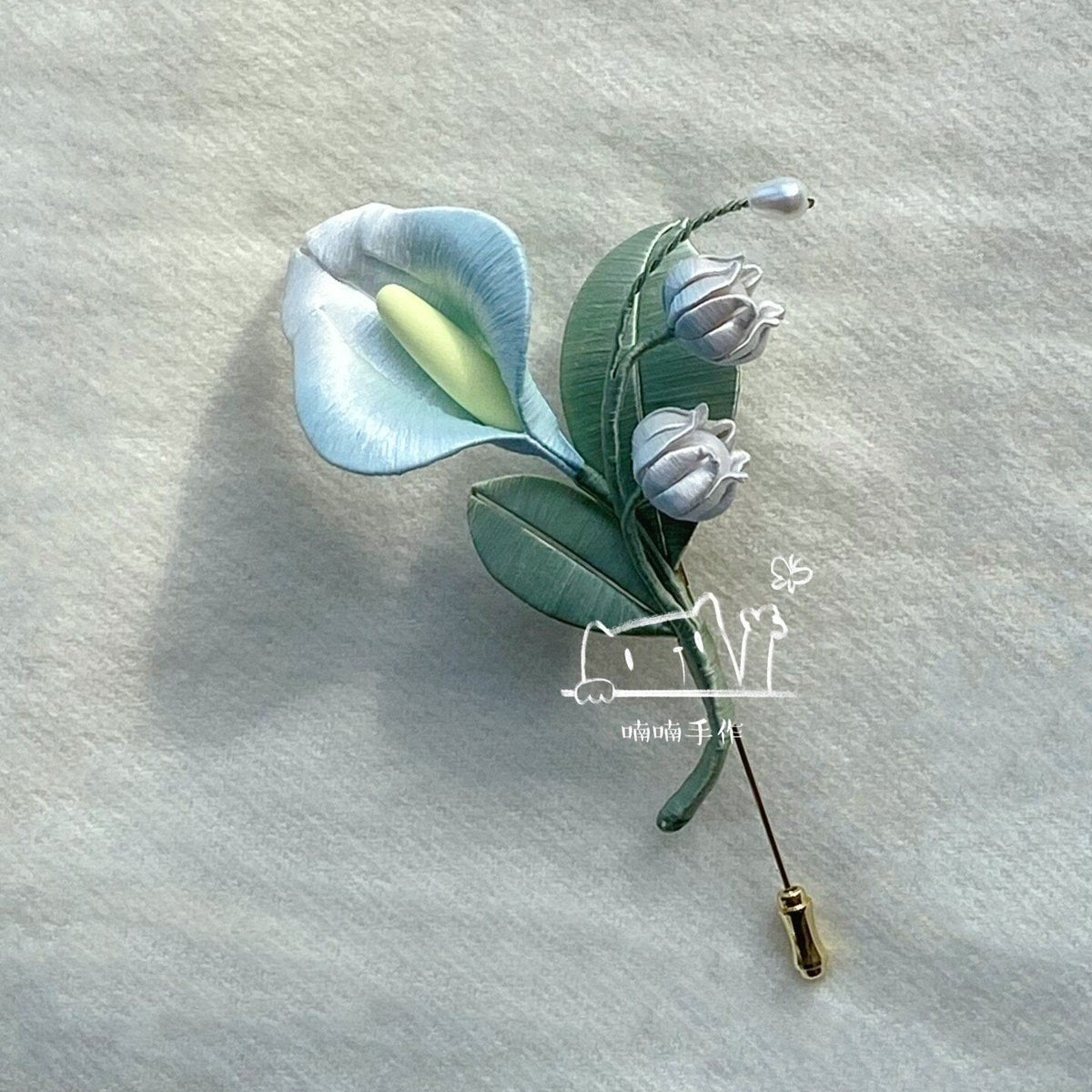 Handmade Chanhua brooch with lifelike light blue calla lily and white lily of the valley displayed on soft off-white fabric