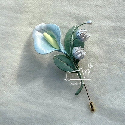 Handmade Chanhua brooch with lifelike light blue calla lily and white lily of the valley displayed on soft off-white fabric