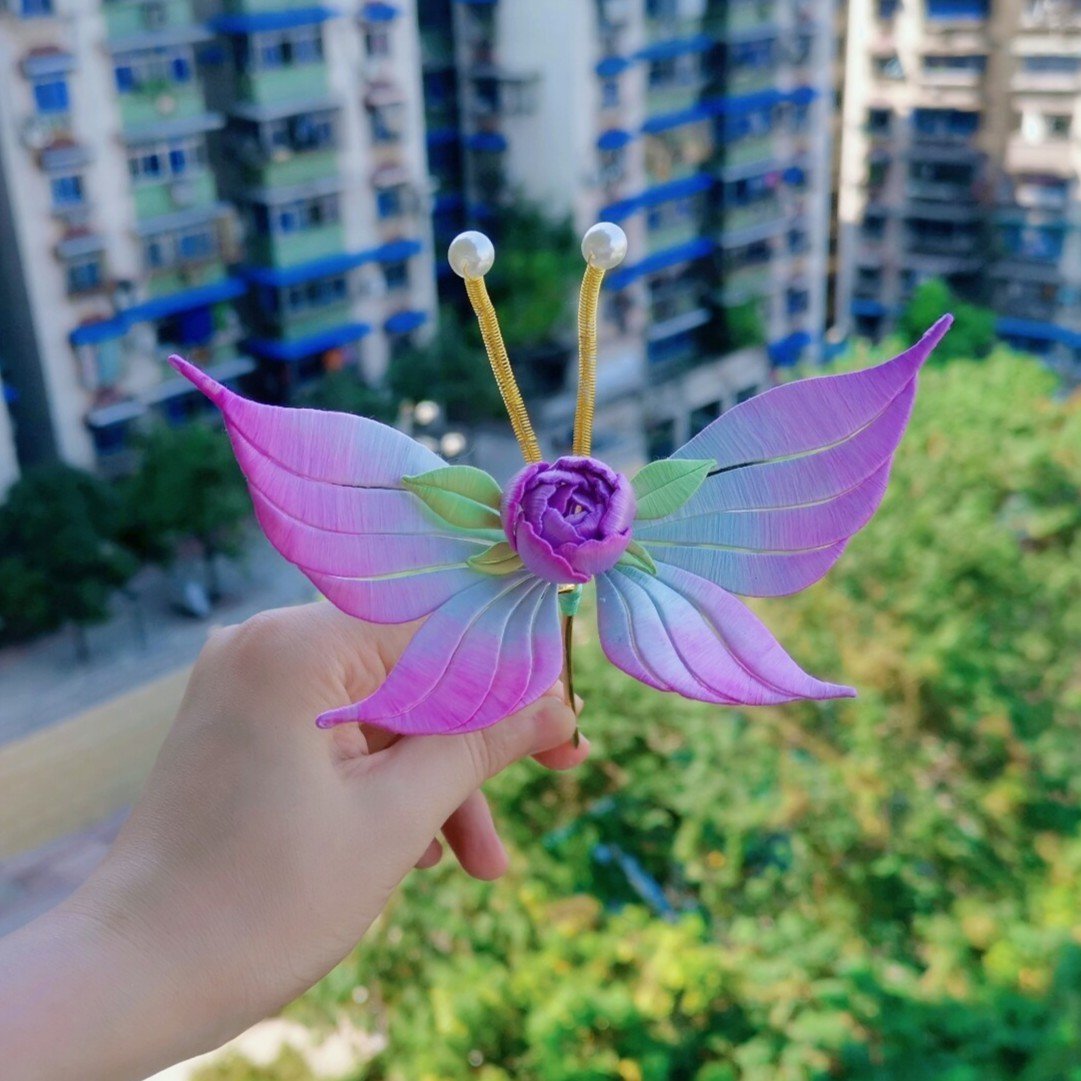 Hand held handmade Chanhua purple gradient butterfly hair accessory against outdoor greenery and residential buildings background