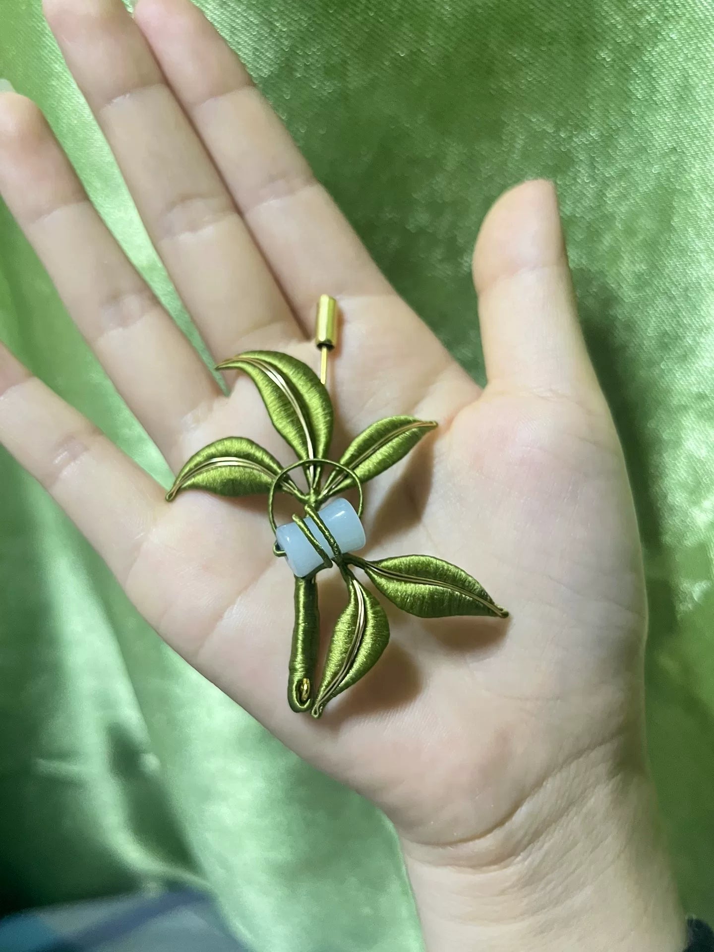 A handmade Chanhua bamboo leaf brooch with green leaves and light blue beads held in a hand