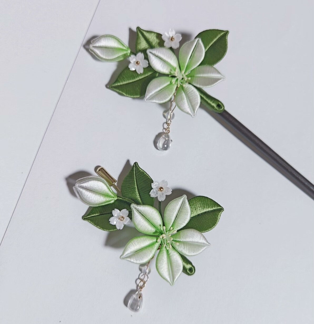 Chanhua handmade jasmine hairpin with white and green flower, dark green leaves, small white buds, and crystal drop pendant, displayed with dark metal pin on white background