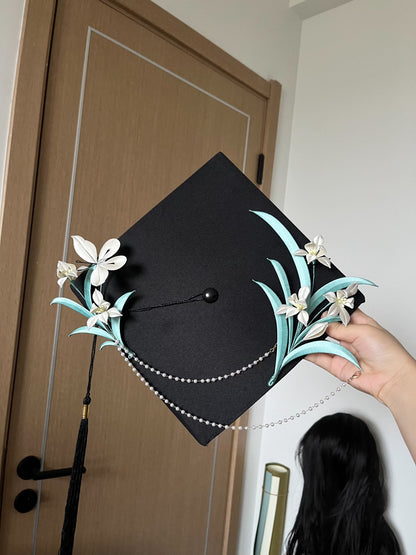 A black graduation cap adorned with white handmade Chanhua and turquoise - colored leaves, held by a hand