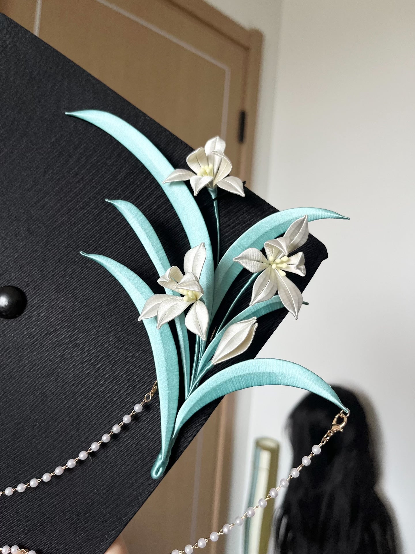 A black graduation cap adorned with white handmade Chanhua and turquoise - colored leaves, held by a hand-3