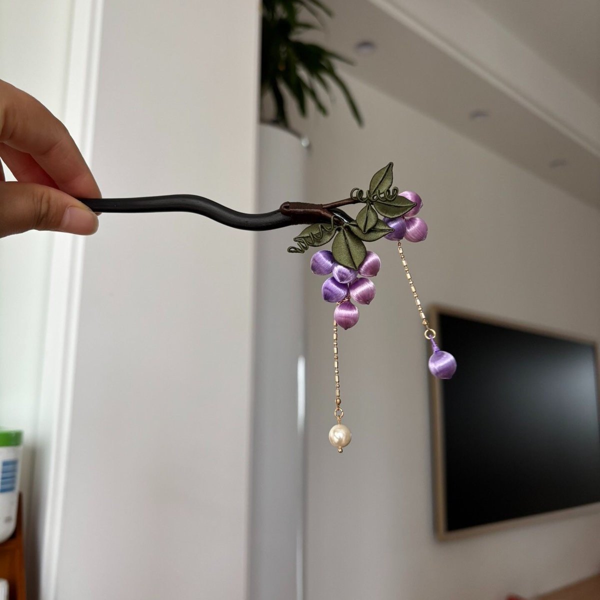 Handheld view of a handcrafted Chanhua thread-wrapped hairpin featuring a cluster of purple grapes, green leaves, and pearl tassel drops.