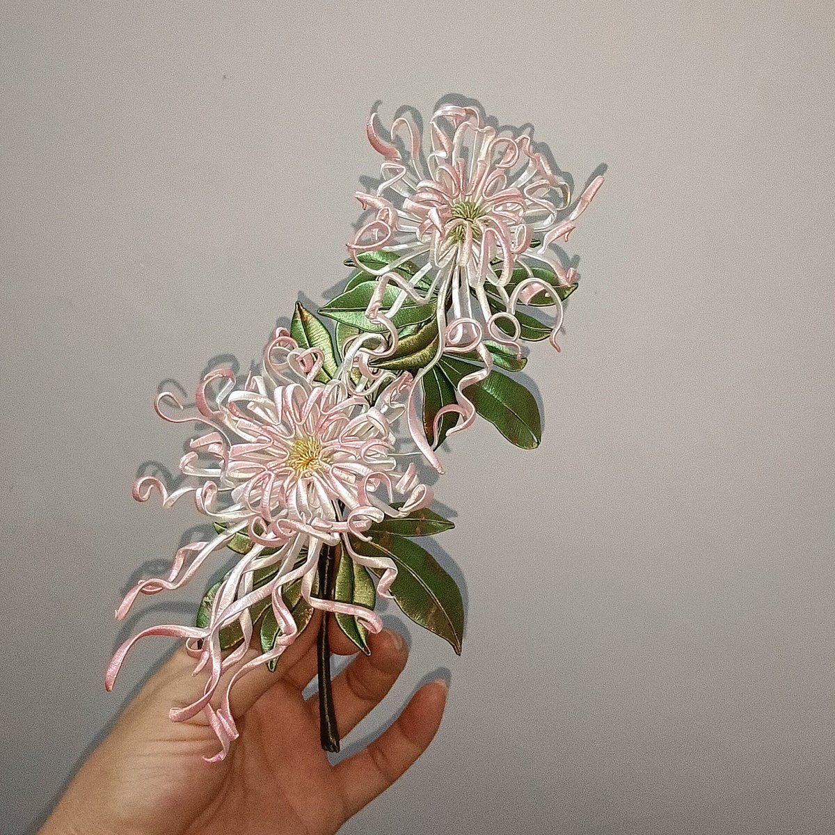 Handheld view of a large, handcrafted Chanhua thread-wrapped pink spider chrysanthemum ornament with two blooming heads and green leaves.