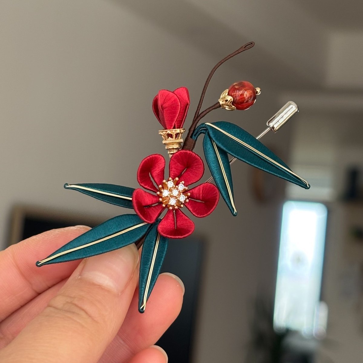 Chanhua handmade plum blossom and bamboo brooch with red flower, crystal stamen, teal gold-edge bamboo leaves, red lacquer stone bead, and dark wire branch, held in hand against a grey background