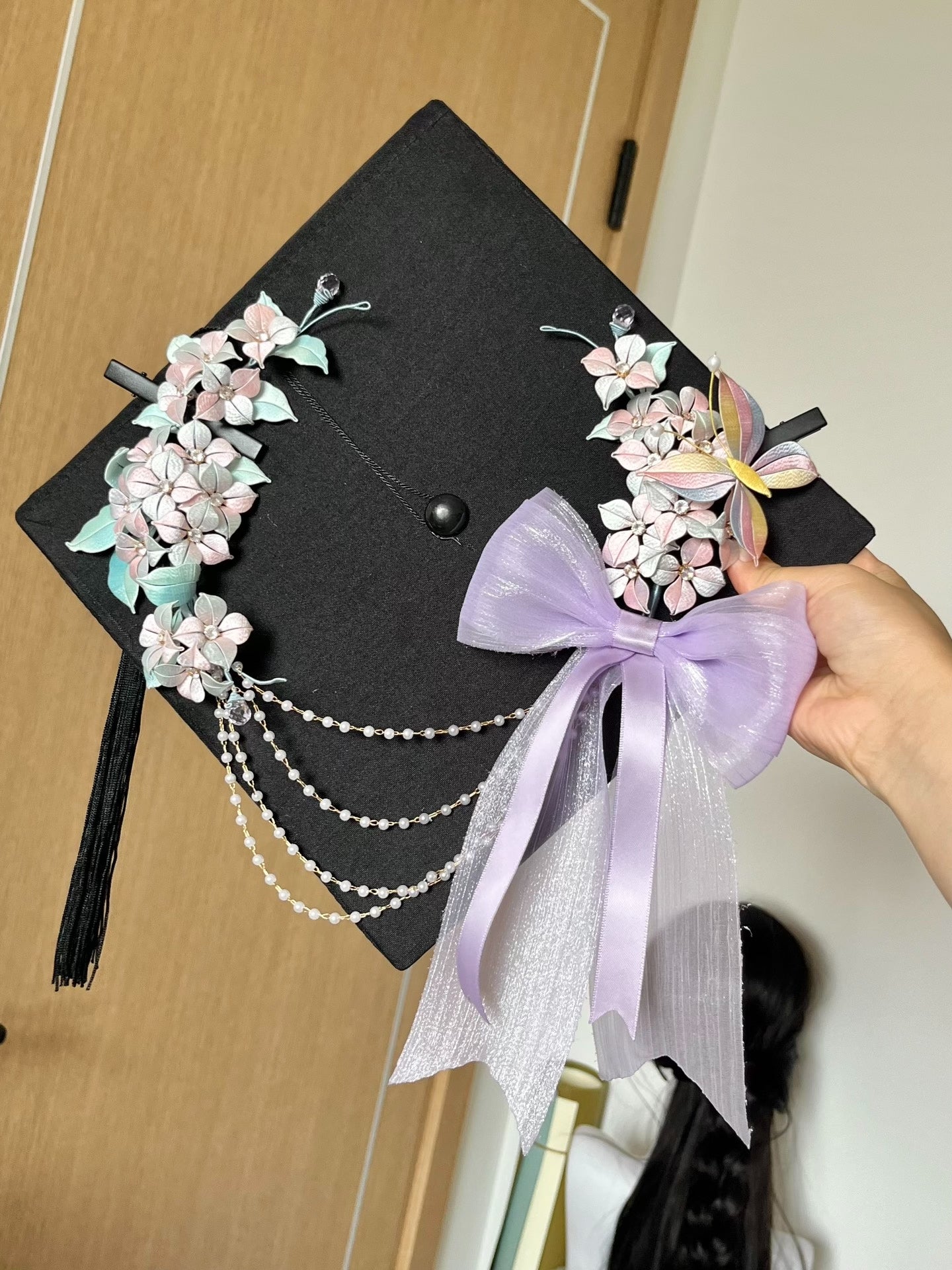 A black graduation cap adorned with handmade Chanhua hydrangea, butterfly, and a lavender bow