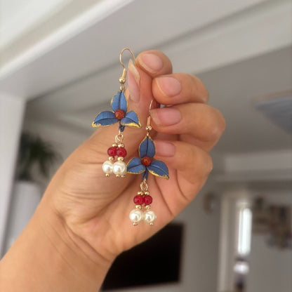 Handheld view of a pair of handmade Chanhua thread-wrapped earrings featuring vintage blue petals with gold edges, red beads, and pearl drops.