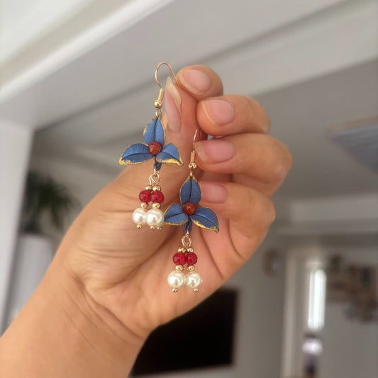Handheld view of a pair of handmade Chanhua thread-wrapped earrings featuring vintage blue petals with gold edges, red beads, and pearl drops.