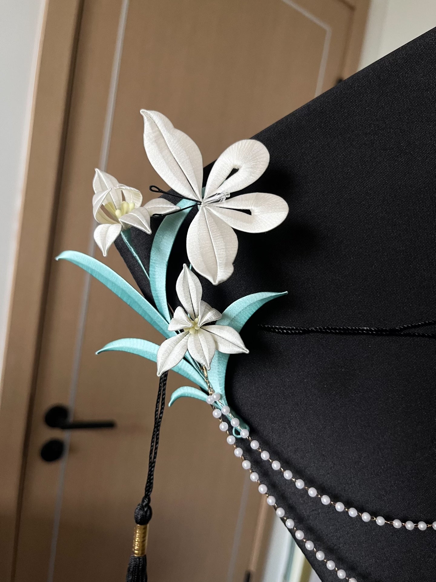 A black graduation cap adorned with white handmade Chanhua and turquoise - colored leaves, held by a hand-2