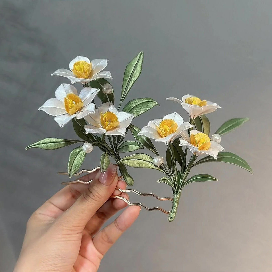 Hand holding a finished white and yellow silk Narcissus Chanhua flower hairpin with green leaves and pearl accents, made from a DIY flower crafting kit.
